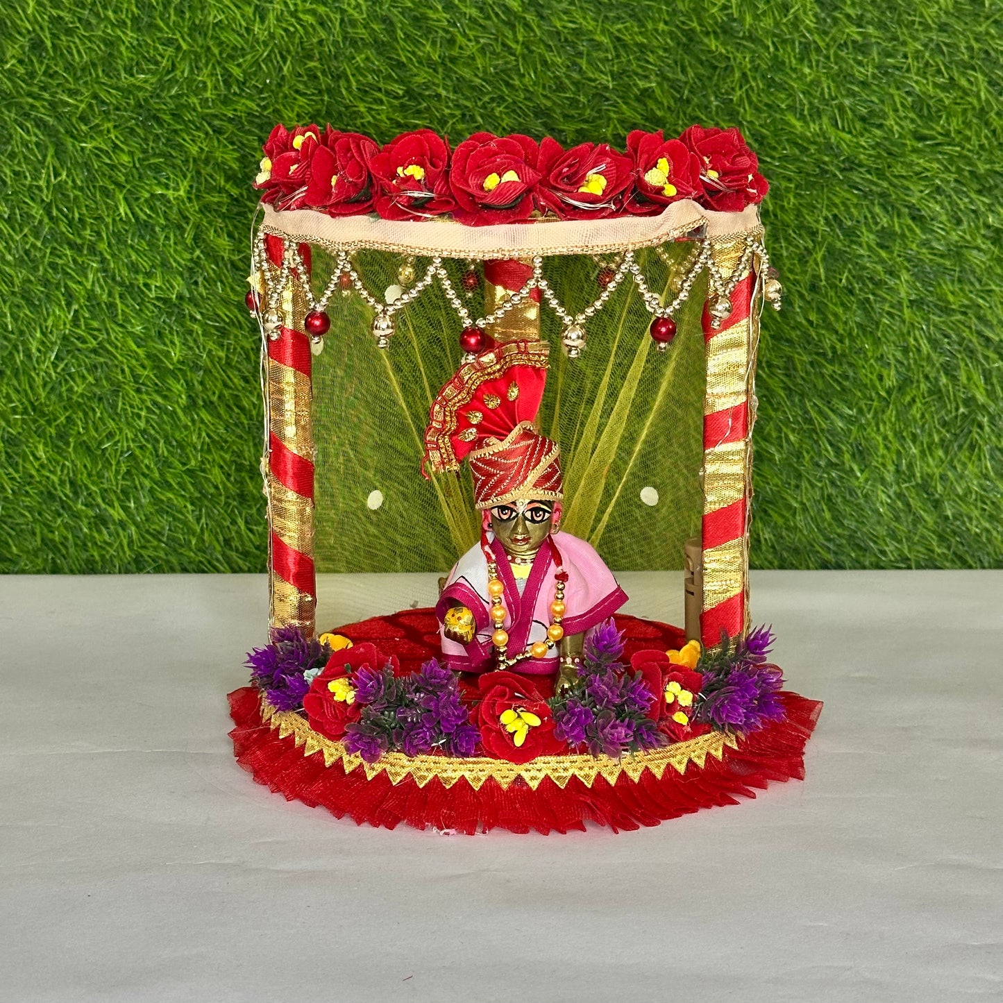 Laddu gopal ji sitting on a decorative Red Floral Laddu Gopal ji Singhasan Bed with Pearl Hangings & LED Lights.