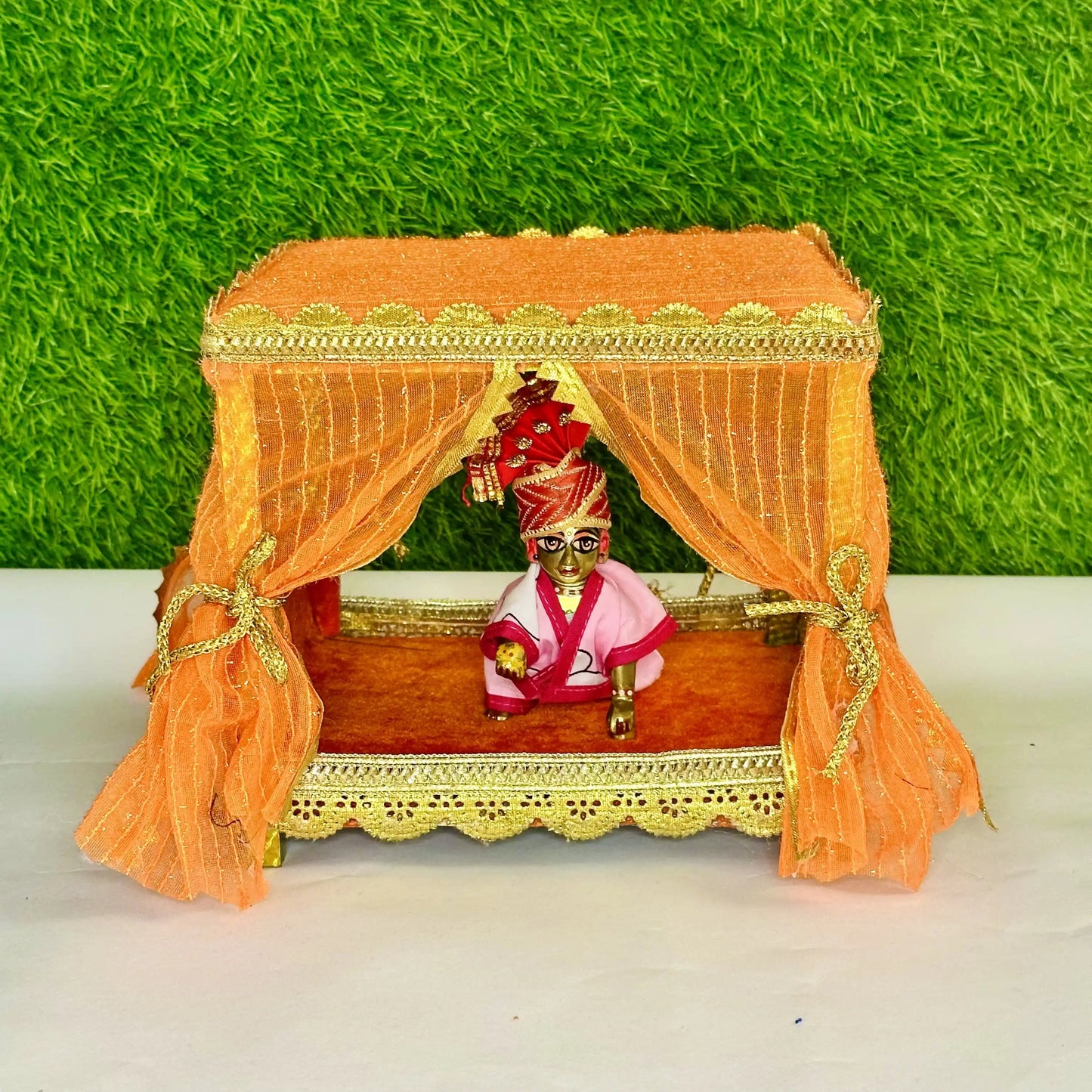 Kanha ji sitting in an Elegant Orange Decorative Laddu Gopal Singhasan with Designer Jhula with Curtains.