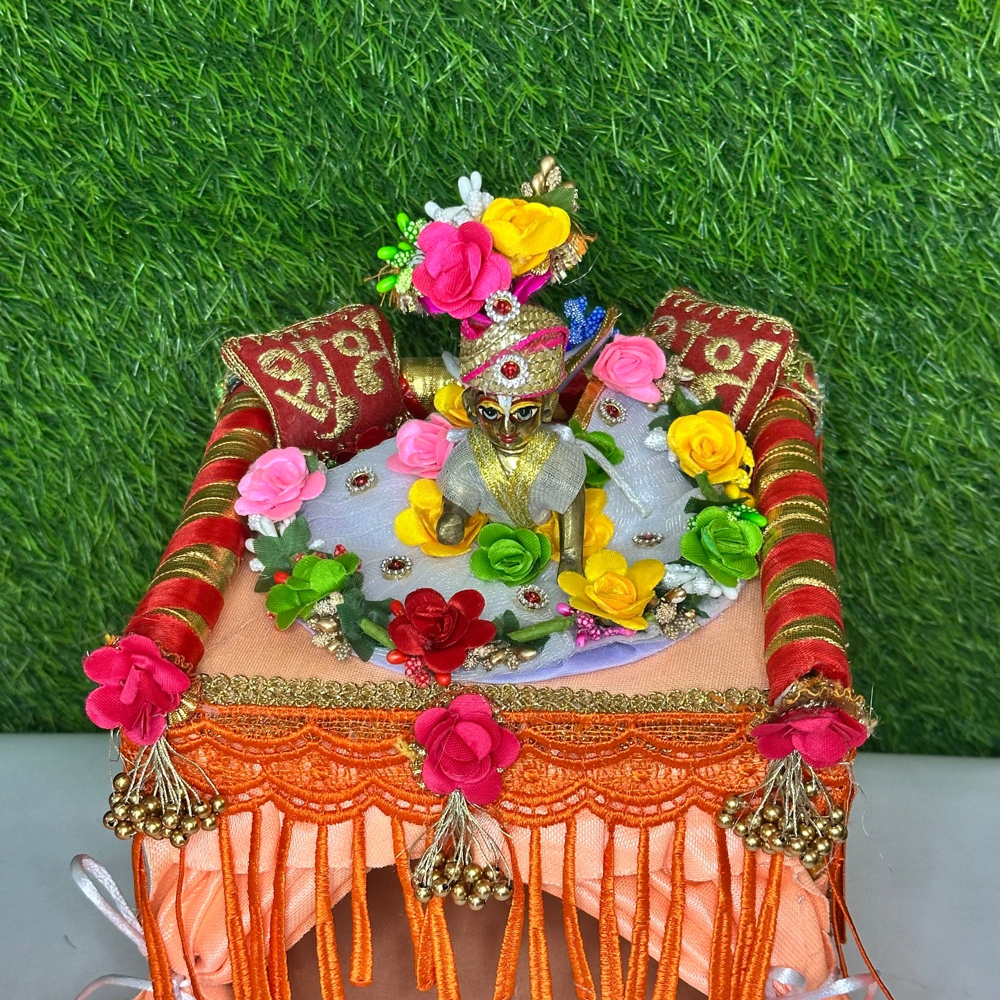 Close up look of laddu gopal ji sitting on a peach color velvet singhasan bed which has decorative canopy, floral pillows & tassel curtains.